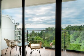 Un balcon agrémenté de deux fauteuils en osier et d’une table en verre, offrant une vue sur un paysage verdoyant et luxuriant ainsi que sur un bâtiment moderne doté de nombreux balcons.