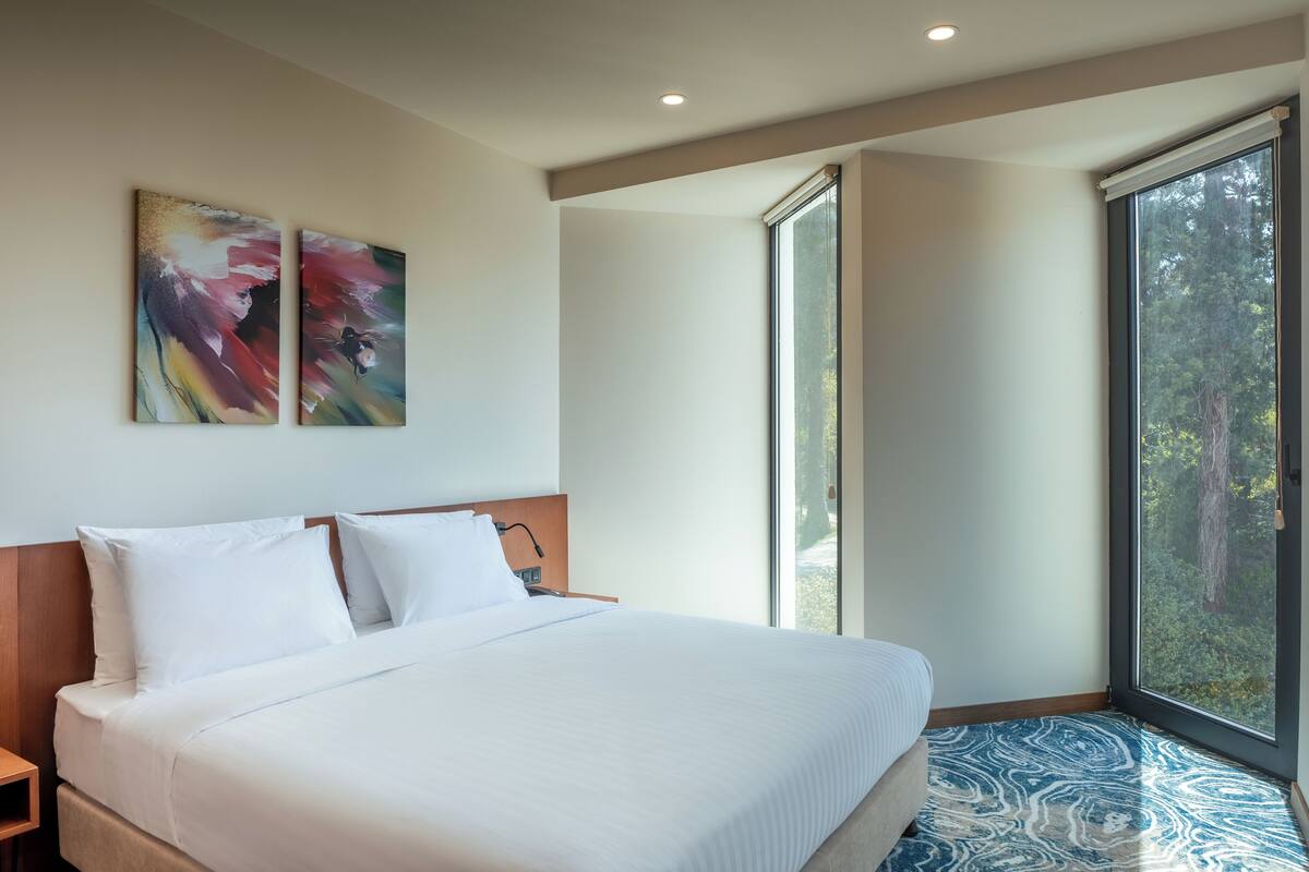 A modern hotel room with a large bed, a nightstand, a window with a view of trees, and abstract art on the wall.