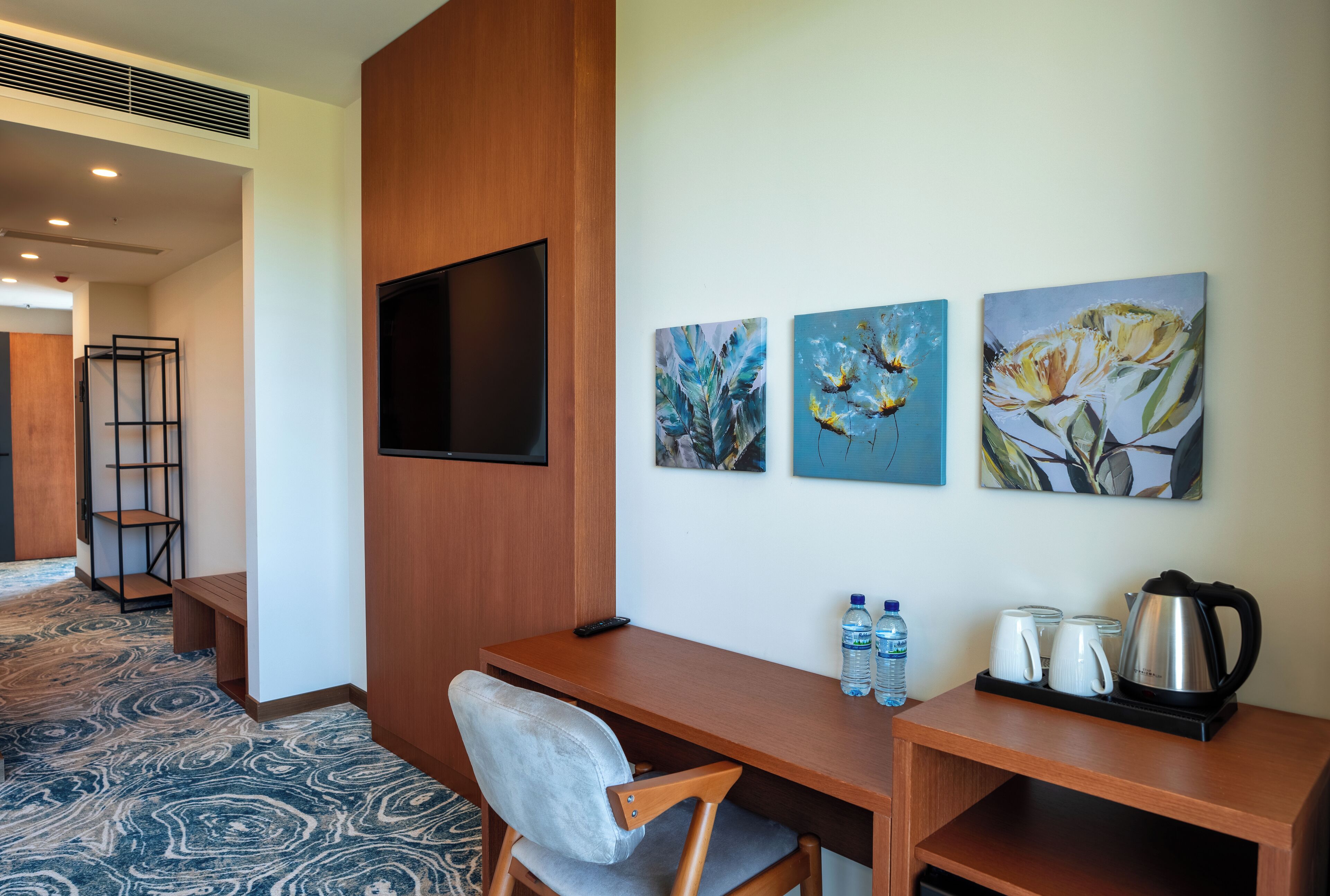 A hotel room with a wooden desk, a chair, a flat-screen TV mounted on the wall, and three framed floral paintings.
