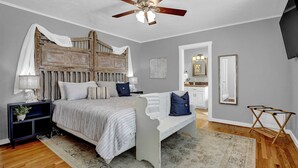 A bedroom with a bed, nightstands, a ceiling fan, a bench, a rug, and a wall-mounted TV. - Bell Haus - Hot Tub, Fire Pit & Walk to Main St (Fredericksburg)