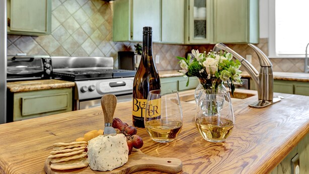 A kitchen counter with a bottle of beer, a glass of wine, cheese, grapes, and a vase with flowers. - Bell Haus - Hot Tub, Fire Pit & Walk to Main St (Fredericksburg)