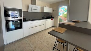 A modern kitchen with white cabinets, a built-in oven, a coffee machine, and a small dining area with a stool. - Villa Avec Piscine,barbecue,terrasse,proche de la Mer. Idéal Pour les Familles (Banyuls-dels-Aspres)