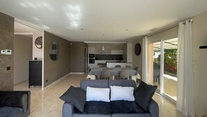 A modern living room with a grey sofa, a dining area with a table and chairs, and a kitchen in the background. - Villa Avec Piscine,barbecue,terrasse,proche de la Mer. Idéal Pour les Familles (Banyuls-dels-Aspres)