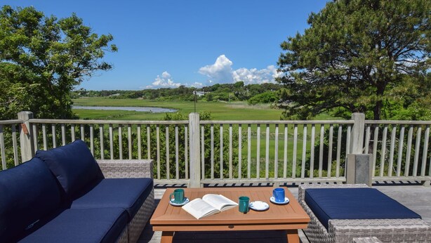 An outdoor seating area with a wooden table, two blue cushioned sofas, and a view of a golf course. - Stunning Marsh Views, Near Pleasant Street Beach! (Chatham)