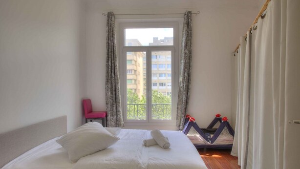 Habitación de hotel con cama, una silla, ventana con cortinas y balcón con vista a edificios.