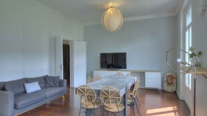 Una sala de estar moderna con un sofá gris, una mesa de comedor con sillas de mimbre, un televisor de pantalla plana y un ventanal grande.
