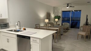 Uma cozinha moderna com lava-louças, forno embutido e uma ilha grande com pia. Há uma área de jantar com mesa e cadeiras, e uma varanda com vista.