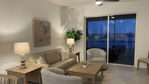 Sala de estar com sofá, poltronas, mesa de centro, abajur, planta e vista para uma varanda com guarda-corpo.