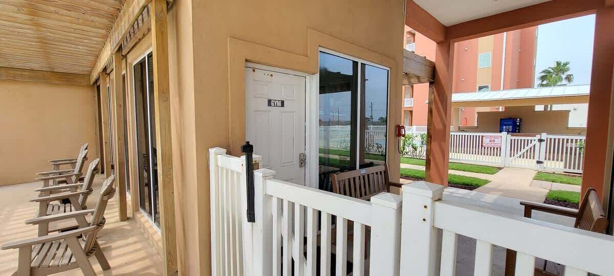 En balkon med gelænder i hvidt, en dør med teksten "GYM" og udsigt til en bygning med rød facade og et hvidt hegn.