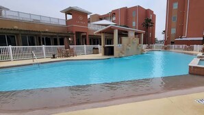 A swimming pool with a white deck, a covered seating area, and a building with balconies in the background. - Like New Killer Patio - Stunner Close to Beach (South Padre Island)