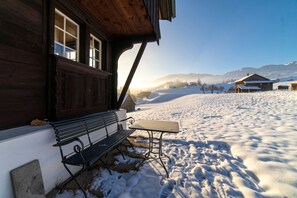 Un edificio de madera con un banco y una mesa en el exterior, rodeado de nieve y montañas al fondo.