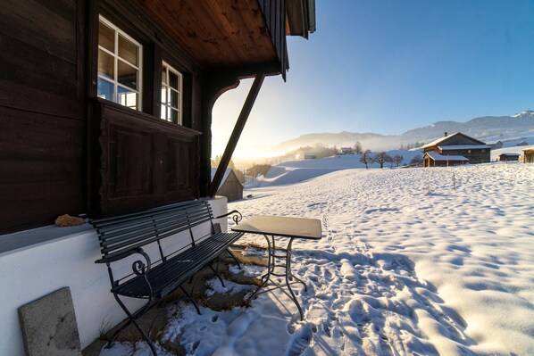 A wooden building with a bench and table outside, surrounded by snow-covered ground and distant mountains.