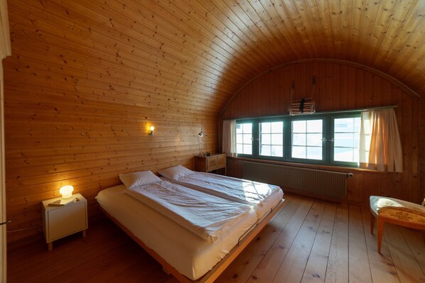 Una habitación acogedora con paredes de madera y techo inclinado, que incluye una cama, lámparas de noche, una ventana con cortinas y un radiador.