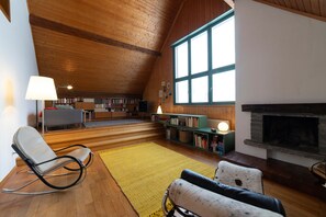 A cozy loft-style living room with a fireplace, bookshelves, a comfortable seating area, and a large window providing natural light.