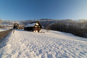 Een houten chalet met een schuin dak, omgeven door een met sneeuw bedekte grond en bomen, onder een heldere lucht.