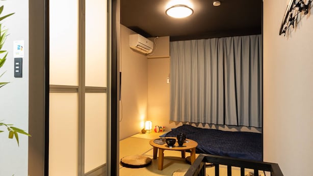 A compact room with a sliding door, a small seating area with a table, and a bed with a blue blanket. - Oyadokinokuniya Isemachi (Nagasaki)
