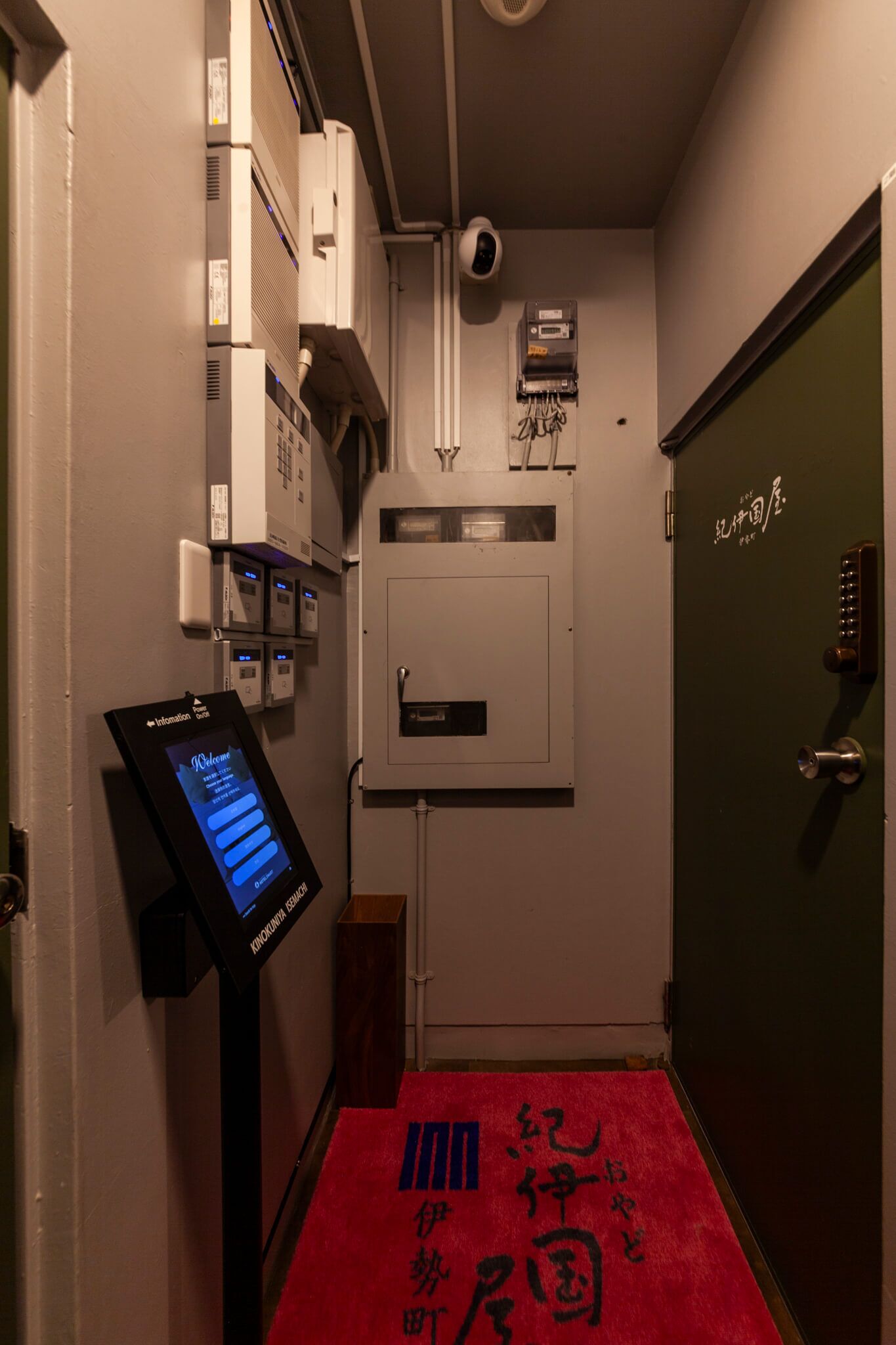 Un pasillo con un quiosco de información digital, una cámara de seguridad y una puerta verde con cerradura.