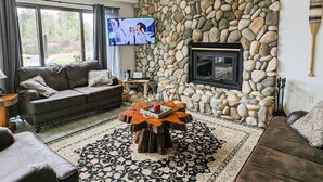 A living room with a stone wall, a fireplace, a television, and a coffee table. - Waterfront close to Traverse City, Kayaks, Row Boat, Firepit, Dog ok, 8pl (Traverse City)