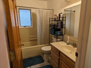 A bathroom with a bathtub, toilet, and sink. There is a window above the bathtub and a shelf with toiletries.