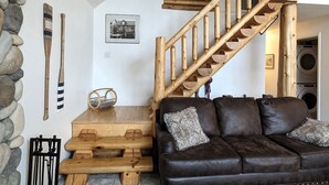 A cozy living room with a stone wall, wooden furniture, a brown leather sofa, and a wooden staircase. - Waterfront close to Traverse City, Kayaks, Row Boat, Firepit, Dog ok, 8pl (Traverse City)