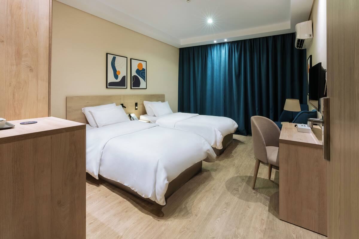A hotel room with two beds, a desk, a chair, a TV, and artwork on the wall.