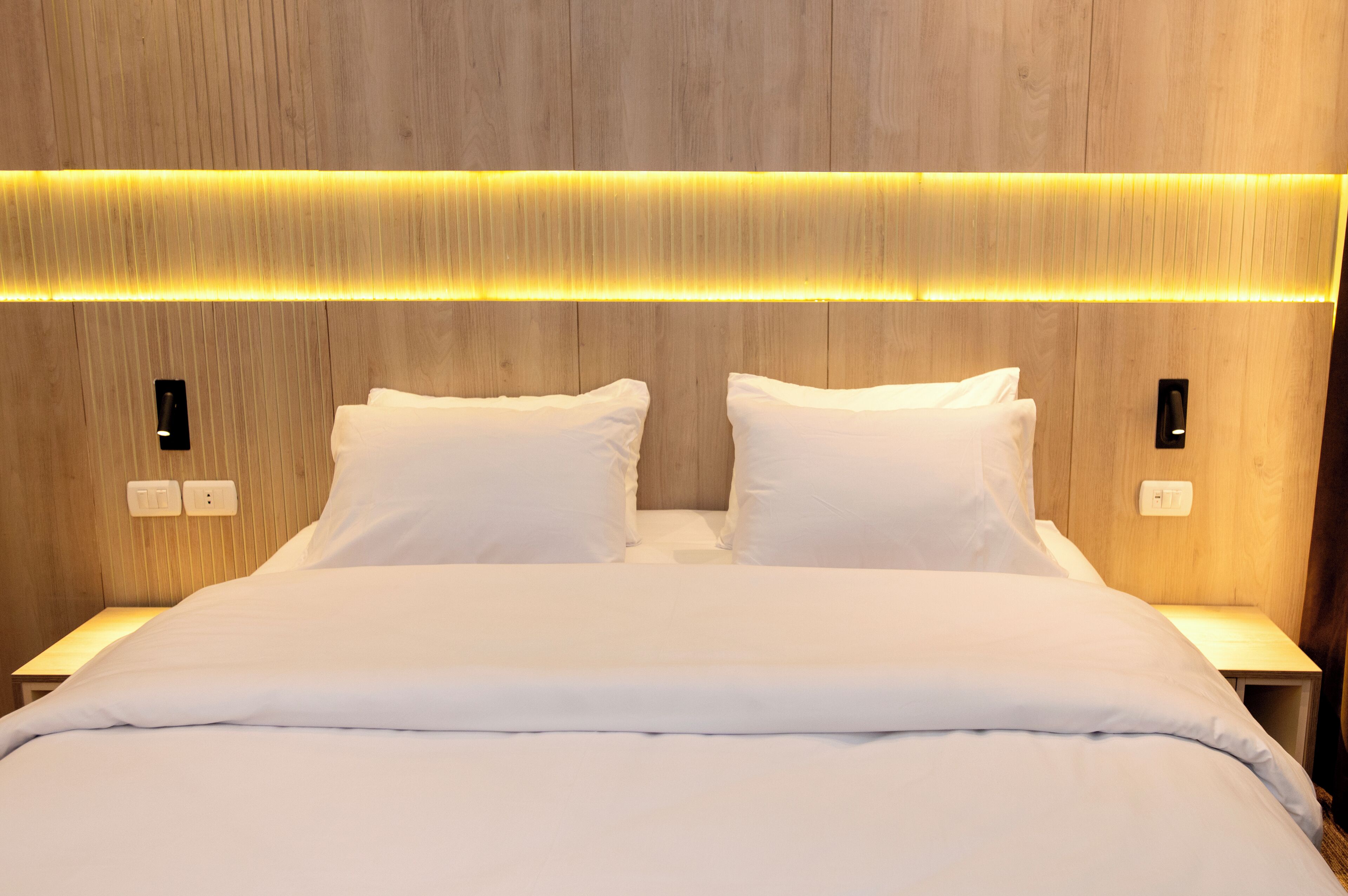Ein ordentlich bezogenes Bett mit weißen Leinen, ein Holzkopfende mit integrierter Beleuchtung und Nachttische mit Schaltern.