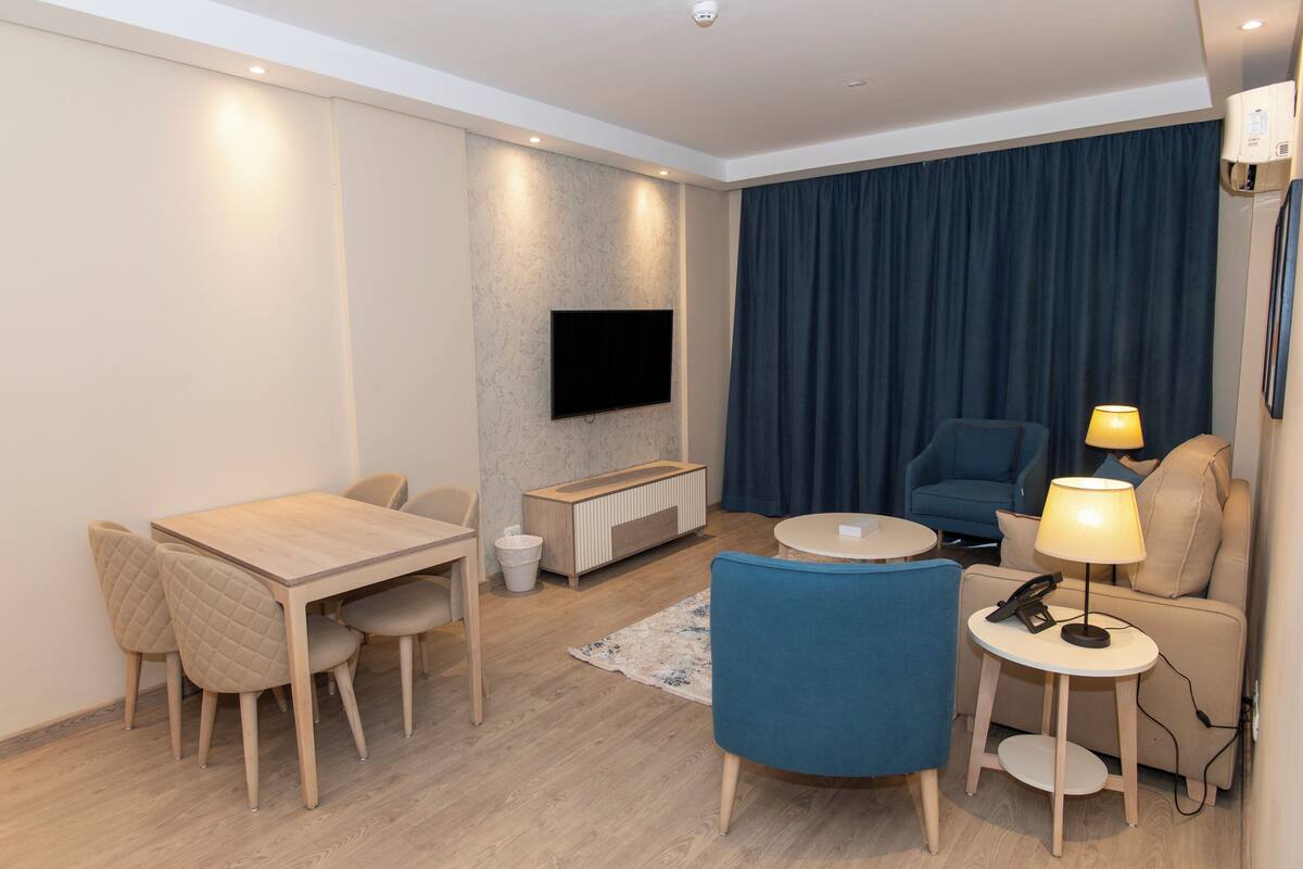 A modern hotel room with a wooden dining table, blue armchairs, a sofa, a TV mounted on the wall, and a small side table with a lamp.