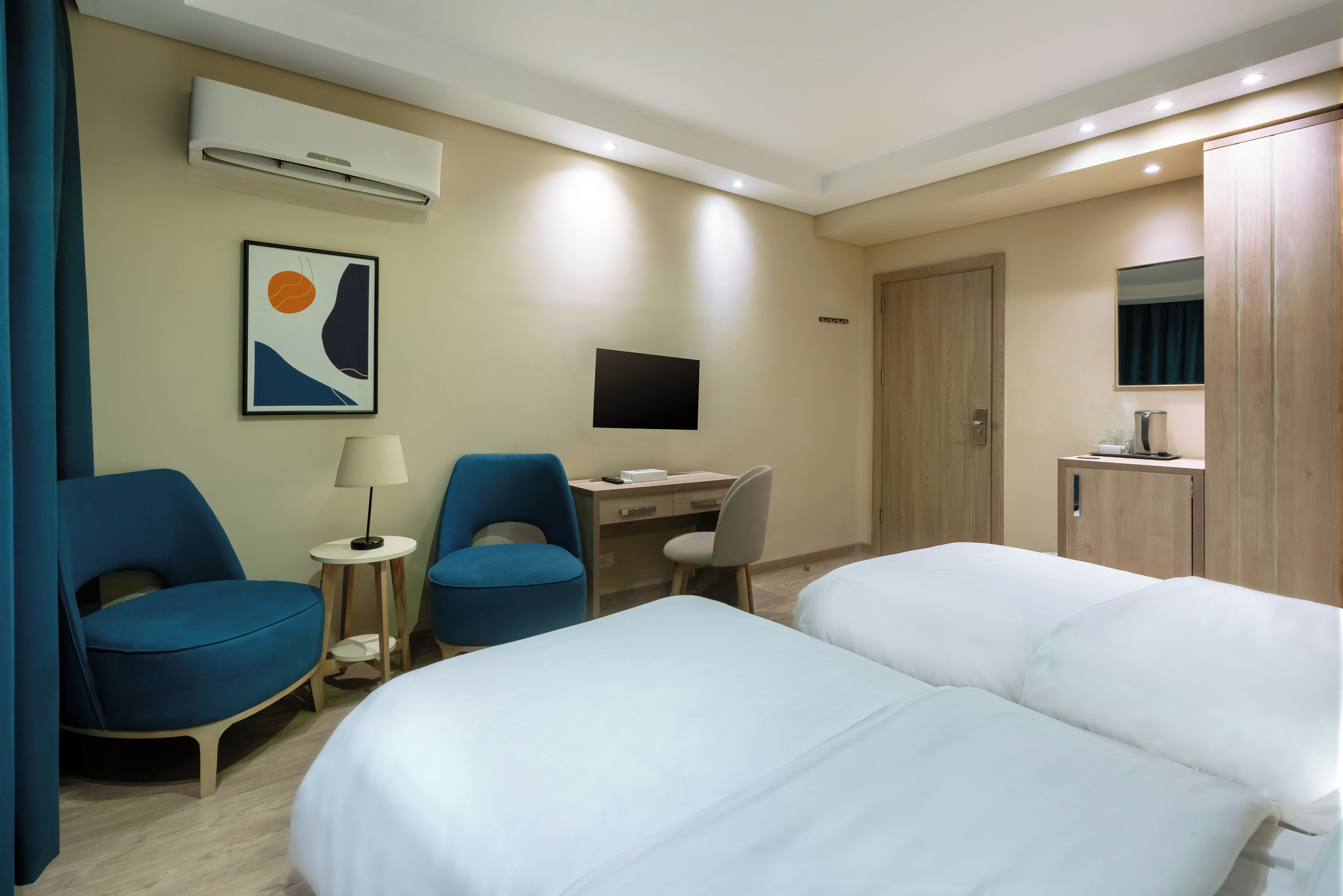 Quarto de hotel com duas camas, uma escrivaninha com computador, televisão e guarda-roupa.