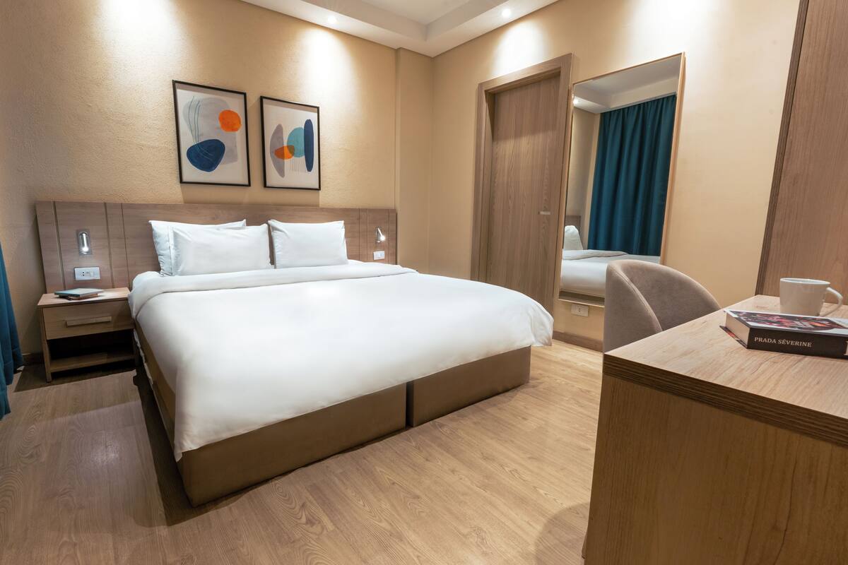 A hotel room with a large bed, a desk with a book, a mirror, and two abstract paintings on the wall.