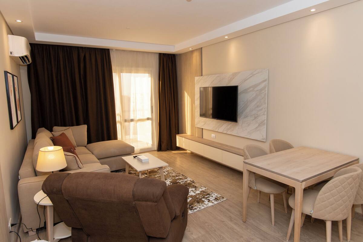 A modern living room with a sofa, a coffee table, a TV mounted on the wall, and a dining area with a table and chairs.