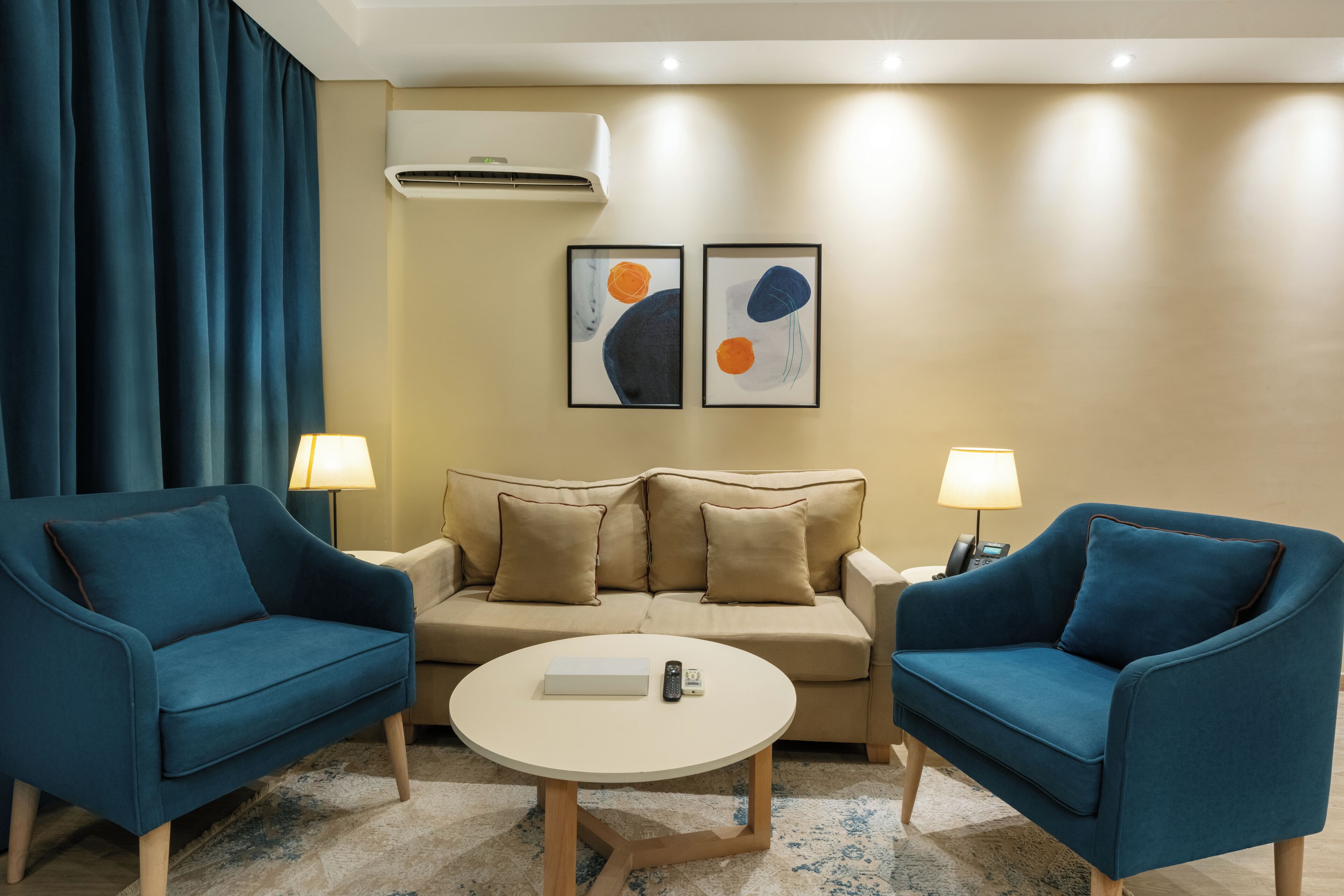 Ein modernes Hotelzimmer mit einem beigen Sofa, zwei blauen Sesseln, einem runden Couchtisch und zwei abstrakten Bildern an der Wand.