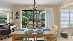 Un coin repas avec une table en bois, des chaises et un lustre. Il y a deux fenêtres équipées de stores, un canapé avec des coussins et une vue sur un jardin et des bâtiments à l’extérieur.