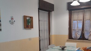 A dining area with a table, chairs, a window with curtains, a ceiling light fixture, and a door with a curtain. - Intero appartamento 70mq  (Scalea)