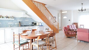 Una cocina con escalera de madera, un comedor con sillas y una mesa para cuatro, y un sillón rosa en la sala de estar.