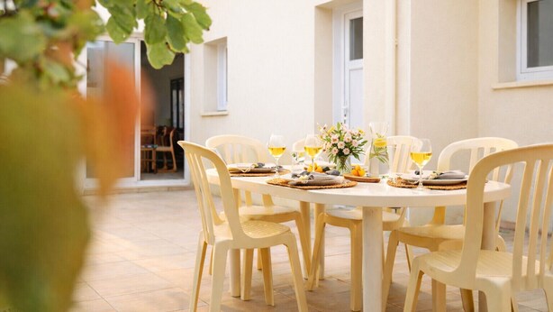 Una mesa redonda al aire libre con sillas blancas, un florero con flores y copas de vino blanco.