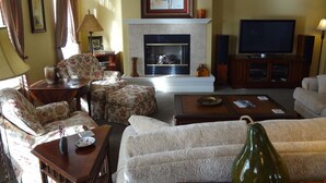 A living room with a fireplace, floral-patterned furniture, a television, and a large window with curtains.