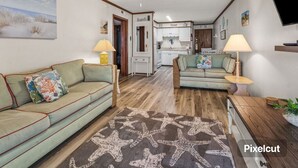 A cozy living room with two sofas, a starfish-patterned rug, and a wooden coffee table. A ceiling fan and a kitchen with white cabinets are visible in the background. -  Cherry Grove Beach Oceanfront 
Finestere -F1 (North Myrtle Beach)