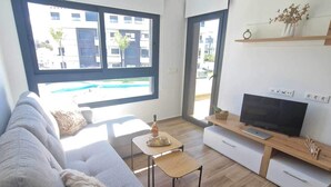 Un salon moderne avec un canapé gris, une table basse, un meuble pour la télévision et une vue sur un immeuble et une piscine à l’extérieur.