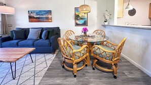 Uma sala de estar com um sofá azul, uma mesa de jantar de vidro cercada por cadeiras de vime, um lustre e um quadro na parede.