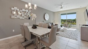 Un espace de salle à manger moderne avec une table blanche, des chaises beiges et un lustre. Une grande fenêtre offre une vue sur la verdure, et un balcon est également présent.