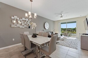 A modern dining area with a white table, beige chairs, and a chandelier. There is a large window with a view of greenery and a balcony.