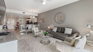 A modern living room with a grey sofa, white armchairs, and a glass coffee table. There is a dining area in the background with a kitchen visible.
