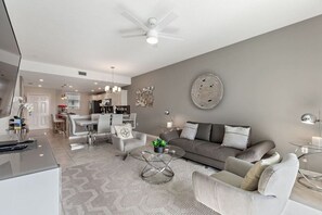Ein modernes Wohnzimmer mit einem grauen Sofa, weißen Sesseln und einem Glastisch. Im Hintergrund ist ein Essbereich mit einer sichtbaren Küche zu sehen.