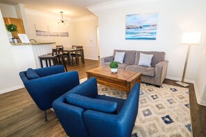 Una sala de estar con un sofá, dos sillones, una mesa de centro y una alfombra. Se observa un comedor con una mesa y sillas al fondo.