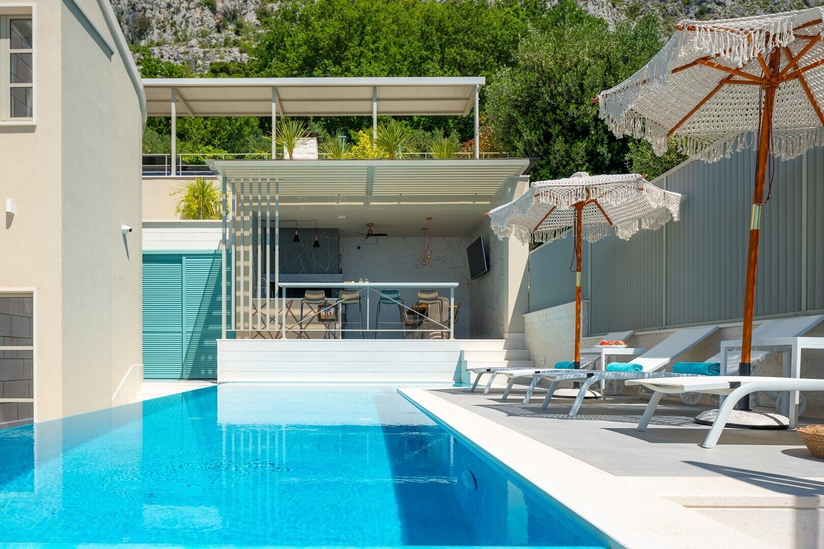 Un espace piscine moderne avec une piscine à l’eau d’un bleu limpide, des chaises longues blanches et un coin salon abrité, équipé d’une table et de chaises.