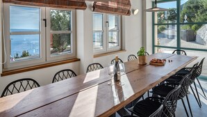 A dining area with a long wooden table, black chairs, and large windows offering views of the sea and greenery. - Casa GENI with a heated pool, 3 bedrooms, an organic garden, sea views (Borak)