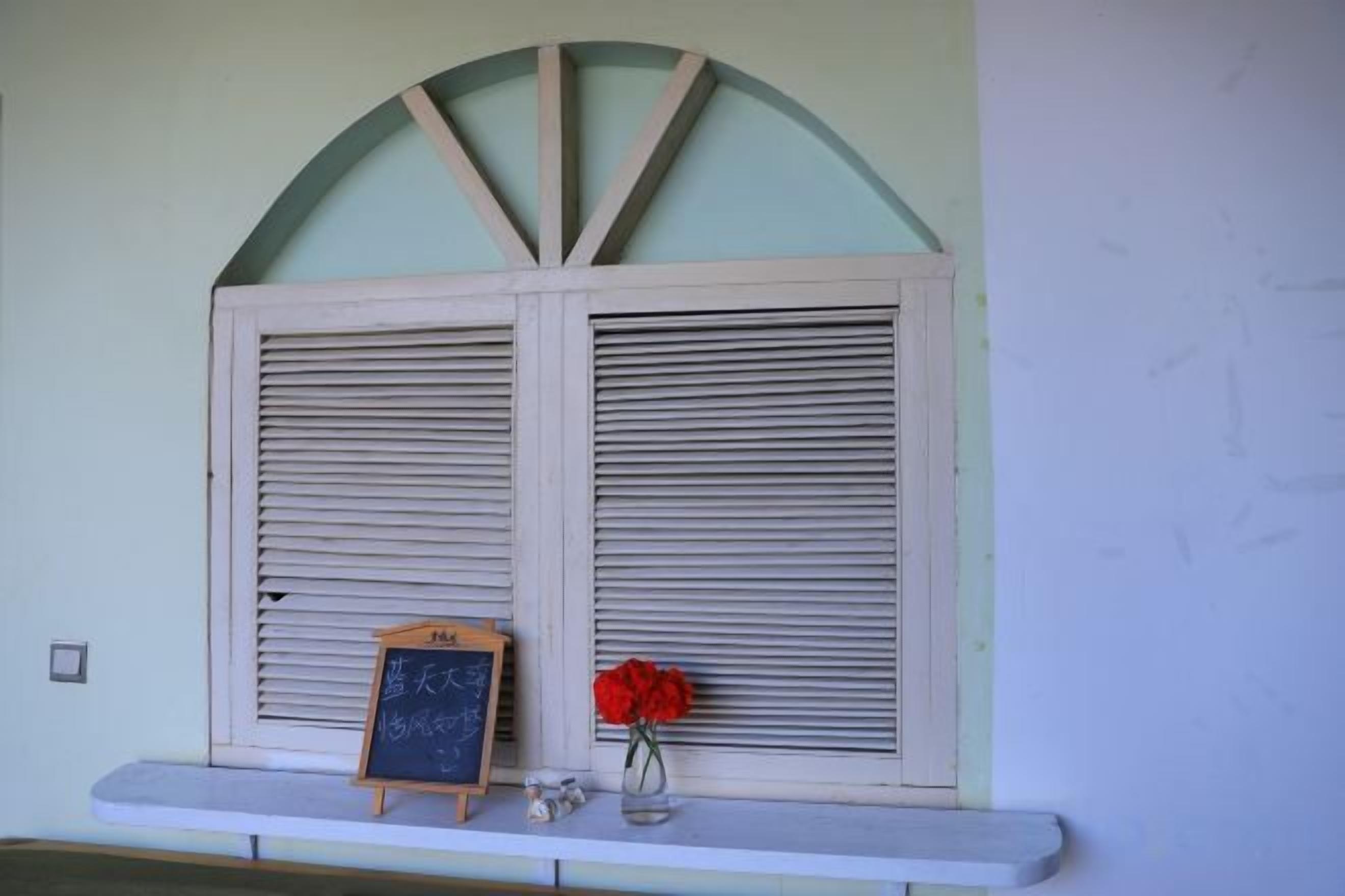A window with shutters, a chalkboard with writing, and a vase with a red flower on a ledge.