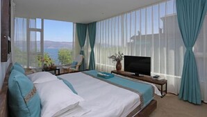 Un dormitorio con una cama grande, un televisor sobre un soporte de madera y vista al mar a través de cortinas translúcidas.