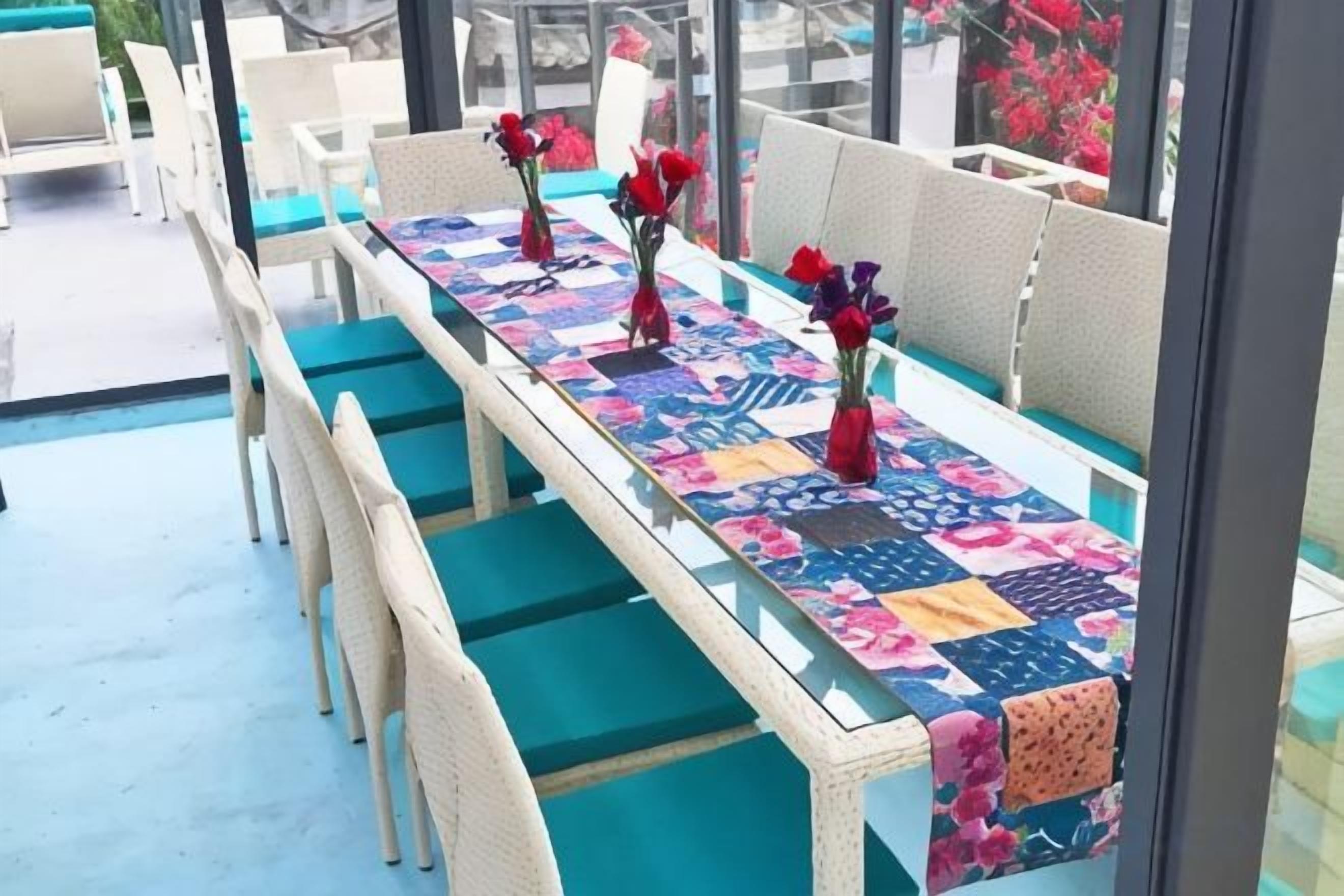 A long dining table with chairs, a patterned tablecloth, and red flowers in vases.
