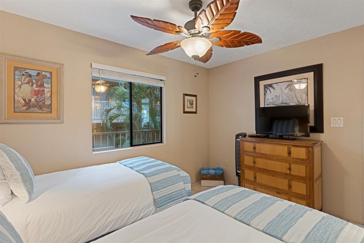 A room with two beds, a ceiling fan, a TV, a dresser, and a window with a view of palm trees.